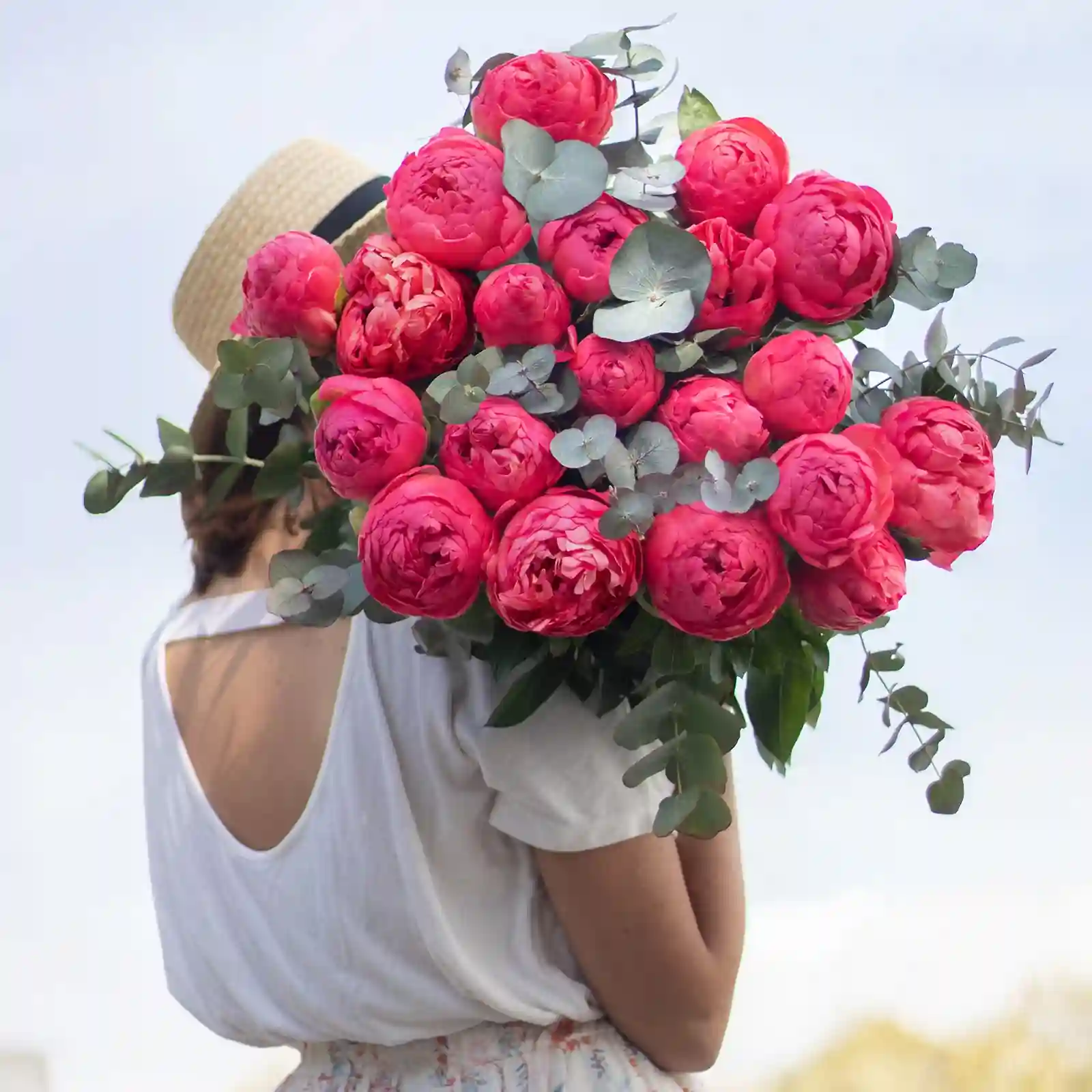 The image shows a person from behind holding a large bouquet of vibrant pink peonies mixed with eucalyptus leaves. The person is wearing a white shirt with an open back and a straw hat. The background is a clear sky, suggesting an outdoor setting. The bright colors of the flowers contrast beautifully with the person's attire and the sky, making the bouquet the focal point of the image.