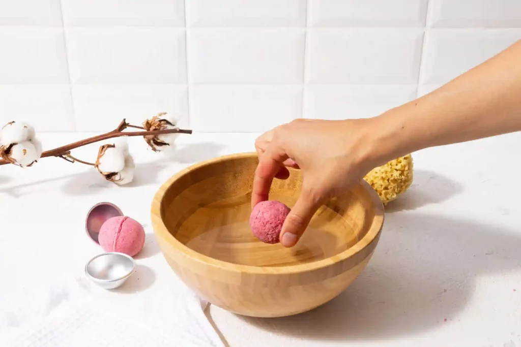 The image shows a hand holding a pink bath bomb over a wooden bowl. There is another pink bath bomb in a metal mold next to the bowl. In the background, there is a branch with cotton bolls and a natural sponge. The setting appears to be a clean, white surface with a tiled wall in the background. The image seems to be related to self-care or bath products, highlighting the use of natural and handmade items. Bath bombs are popular for creating a relaxing and aromatic bath experience, often with added benefits like moisturizing the skin or providing soothing scents.
