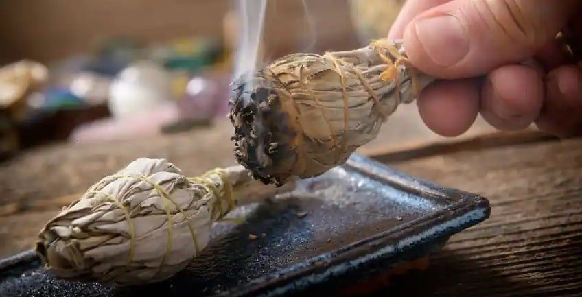 The image shows a hand holding a burning bundle of sage, which is emitting smoke. Another bundle of sage is placed on a ceramic dish or tray. The background includes blurred objects, possibly crystals or stones, suggesting a setting related to spiritual or cleansing practices. This image is interesting and relevant as it depicts the practice of smudging, which is often used for purification, cleansing, or ritualistic purposes. Smudging is a ritual that has been part of many cultures for centuries. Sage, known for its purifying properties, is commonly used in this practice to cleanse spaces, objects, or individuals of negative energy.