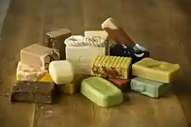 The image showcases a delightful assortment of handmade soap bars, artfully arranged on a rustic wooden surface. These soaps exhibit a variety of shapes, sizes, and colors, reflecting the diverse ingredients and scents they might possess. Some of the bars have textured surfaces, while others are smooth. You’ll notice a mix of solid colors and captivating marbled patterns. Interestingly, one of the soaps features a rope, perfect for hanging.