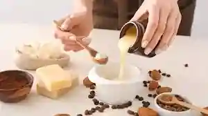 The image shows a person pouring a creamy substance from a jar into a white bowl. The person is holding a wooden spoon with a brown substance, possibly sugar or a spice. Surrounding the bowl are various ingredients, including a block of butter, coffee beans, and small cookies or biscuits. There are also other small bowls containing different ingredients, possibly for a recipe or a skincare preparation. The scene suggests a process of mixing or preparing a recipe, which could be culinary or cosmetic in nature. The image is interesting because it captures a moment of preparation and the use of natural ingredients, highlighting a hands-on, artisanal approach.
