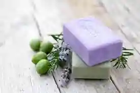 The image shows two bars of soap placed on a wooden surface. One bar of soap is lavender-colored and the other is light green. The lavender-colored soap is placed on top of the green soap. There are also some green olives and sprigs of rosemary and lavender flowers next to the soaps. The scene suggests a natural and possibly handmade or organic theme, emphasizing the use of natural ingredients in the soap.