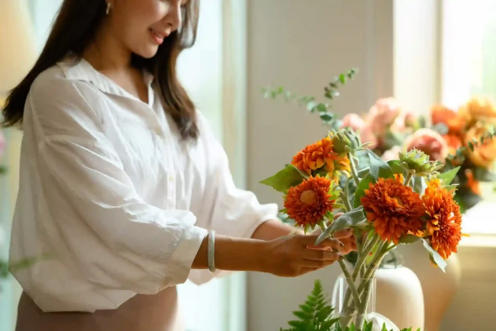 The image shows a person arranging a bouquet of orange flowers in a vase. The person is wearing a white shirt and a bracelet. The background includes a window with natural light coming through and additional flowers in soft focus. The scene appears to be indoors, possibly in a home or a floral shop. The vibrant orange flowers and the act of arranging them make the image visually appealing and relevant for topics related to floristry, home decor, or hobbies involving flower arrangement.