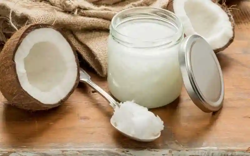 The image shows a jar of coconut oil with its lid off, a spoon with a scoop of coconut oil, and two halves of a coconut. The jar is filled with white, solidified coconut oil. The spoon is placed in front of the jar, and the coconut halves are positioned on either side of the jar. The background includes a piece of burlap fabric and a wooden surface. This image visually represents coconut oil, a popular natural product used for cooking, skincare, and haircare.