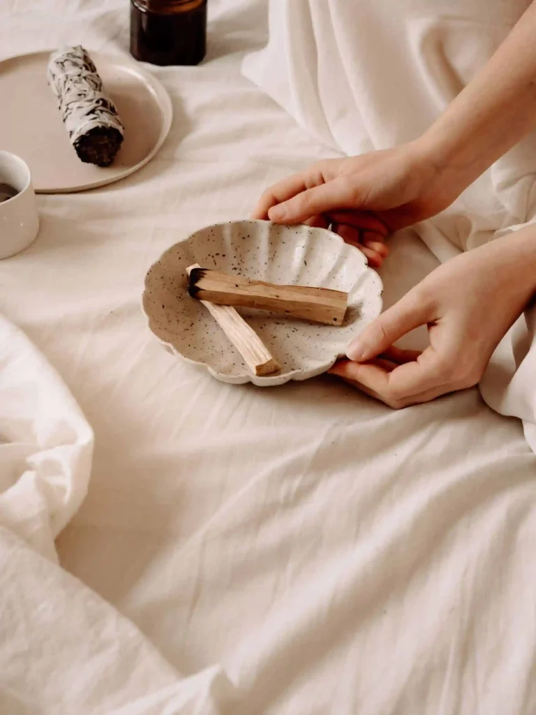 The image shows a pair of hands holding a speckled ceramic dish with two pieces of wood, likely Palo Santo, on a bed with white sheets. To the left, there is a white cup, a plate with a bundle of sage, and a brown bottle. The scene suggests a ritual or practice involving smudging or cleansing, which is interesting as it highlights a cultural or spiritual practice.