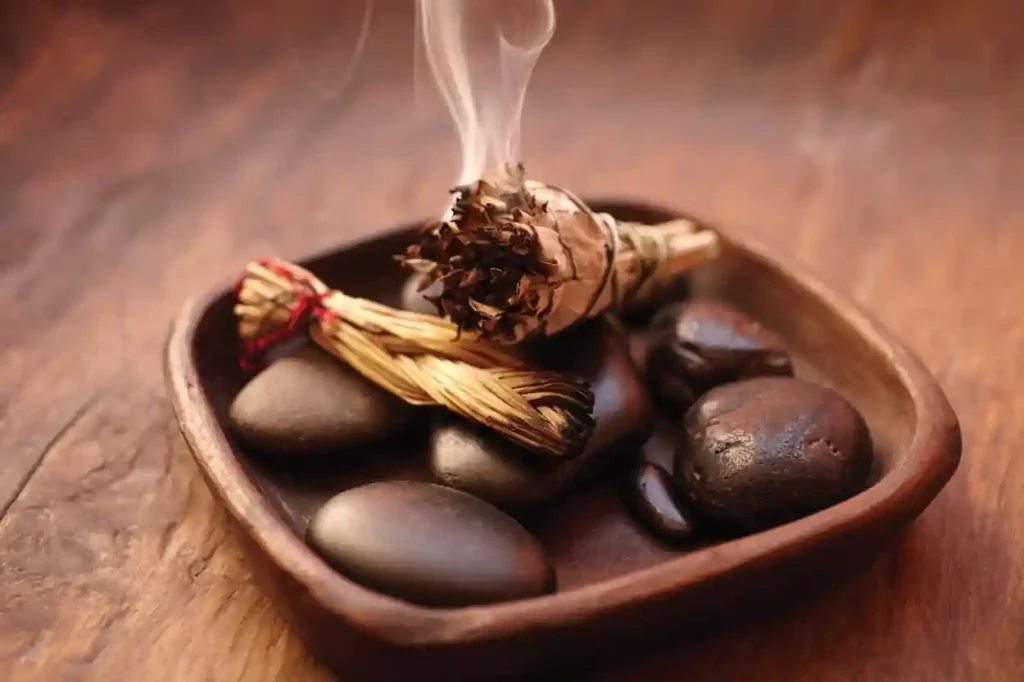 The image shows a wooden tray containing several smooth, dark stones and a bundle of burning sage, which is emitting smoke. There is also a small bundle of straw tied with red string. The scene appears to be set on a wooden surface. This image is interesting and relevant as it depicts items commonly used in practices such as smudging, which is a ritual for cleansing and purifying spaces. The smooth stones may be used for their calming and grounding properties, while the burning sage is traditionally believed to clear negative energy.