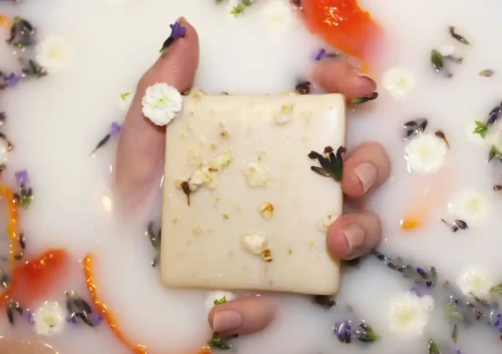 Une main tient un savon rectangulaire dans un liquide laiteux, orné de fleurs séchées, avec des pétales flottants autour.
