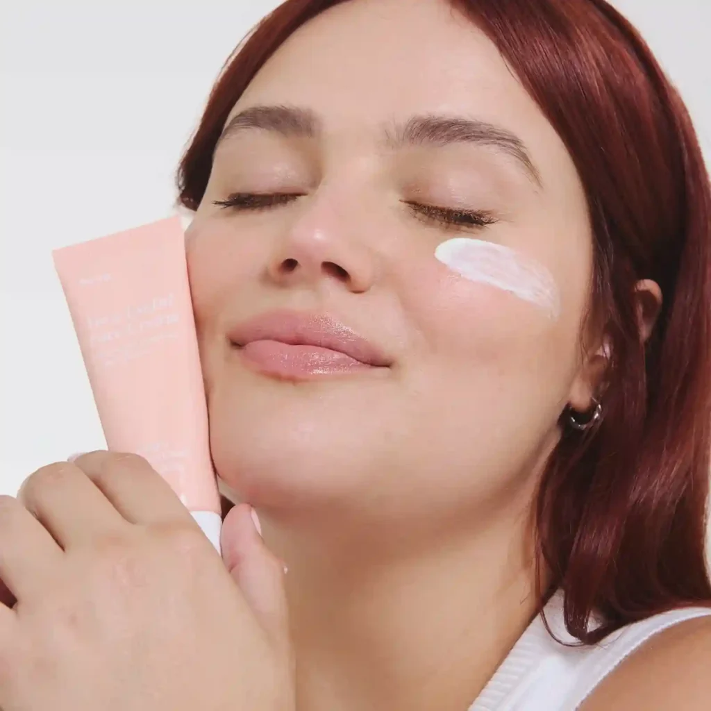 Une personne aux cheveux roux tenant un tube rose de crème pour le visage intitulée « Very Useful Face Cream ».
