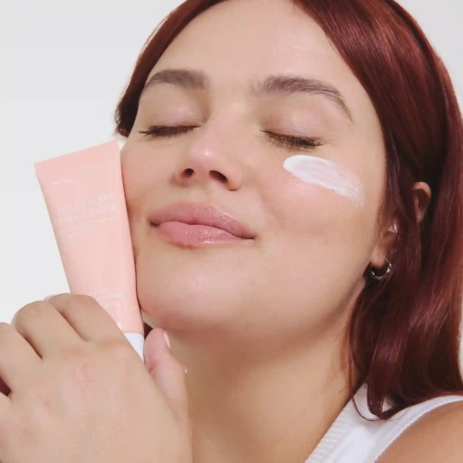 Une personne aux cheveux roux tenant un tube rose de crème pour le visage intitulée « Very Useful Face Cream ».