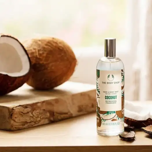 Un flacon de brume pour cheveux et corps à la noix de coco de The Body Shop posé sur une surface en bois, entouré d’un demi-noix de coco et de morceaux de coco.